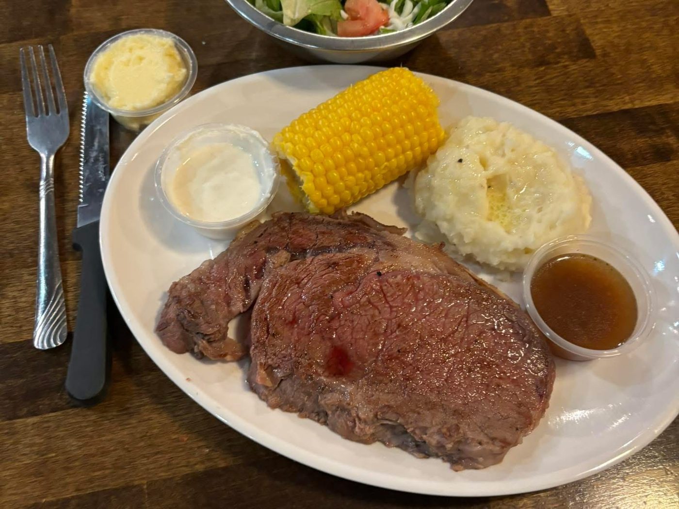 A cut of prime rib with au jus, corn on the cob, mashed potatoes and a side salad